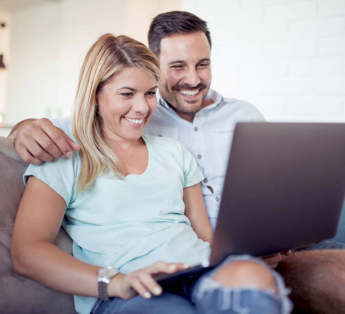 A couple sits closely together on a couch, smiling as they look at a laptop screen, enjoying a moment of connection.