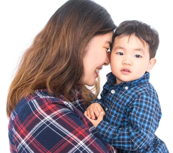 A woman holding a young boy, both wearing plaid shirts, with the woman smiling while the boy gazes thoughtfully.